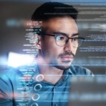 Reputational Damage From Data Breaches and How to Avoid it Ransomware recovery best practices. The image shows a man wearing glasses, intensely focused on a computer screen. He appears to be a programmer or someone working with code, as lines of code and data seem to float in front of him as a digital overlay. The man has short, dark hair, facial hair, and a thoughtful expression as he examines the information. The scene is futuristic, with a soft glow reflecting off the code, evoking a sense of technology and problem-solving, possibly in a cybersecurity or programming environment.