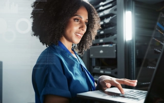 This image shows a person wearing a blue uniform, working with a laptop in what appears to be a server room or data center. The person has curly hair and is focused on the laptop screen while typing. Behind them, there are several server racks and computer equipment, with some light shining from one of the racks, giving a sense of a professional, tech-oriented environment. The person looks engaged and concentrated on their task. Ransomware attack investigation.