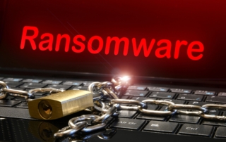 Business continuity plan for ransomware. The image visually represents a ransomware attack. The background shows a laptop keyboard, symbolizing the device being targeted. A heavy metal chain is draped across the keyboard, and a brass padlock secures the chain, symbolizing the device or data being locked or inaccessible. Above the keyboard, in bold, glowing red text, the word "Ransomware" is prominently displayed on a screen, creating a sense of urgency and threat. The red color emphasizes danger, while the chain and lock illustrate the concept of restricted access or encryption often associated with ransomware attacks. The overall tone is one of caution and cyber vulnerability.
