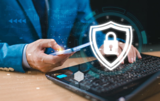SOC team ransomware roles. A person in a blue suit is holding a smartphone in their right hand while their left hand is typing on a black keyboard. There is a digital security overlay on the image, featuring a glowing white shield icon with a padlock in the center, symbolizing cybersecurity. The background is slightly blurred, with a futuristic, technology-inspired theme, including floating digital symbols and circuits.