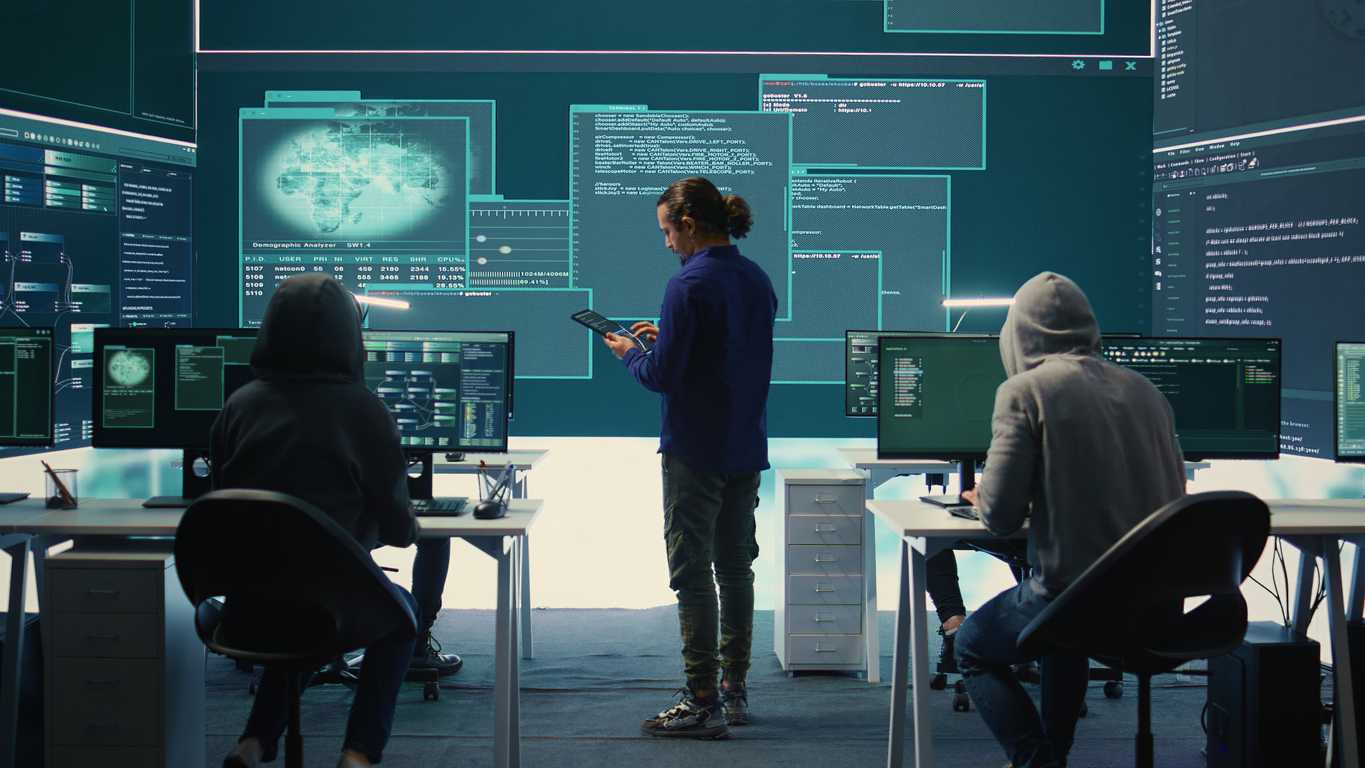Advanced Email Security Protection. Three people are working in a high-tech command center. They face multiple monitors displaying digital blueprints, data streams, and global threat visuals. The middle person stands while using a tablet; the others sit and appear focused on their work. The overall feel is high-intensity and cyber-operations focused.
