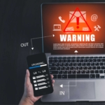 Cloud ransomware vulnerability assessment. A hand holds a smartphone above an open laptop. On the laptop screen is a vivid red warning symbol with an exclamation mark inside a triangle, along with icons for email and settings. White arrows labeled "IN" and "OUT" point between the phone and laptop, suggesting a file upload or data transfer in progress. The scene conveys a cyberattack, security breach, or phishing warning.