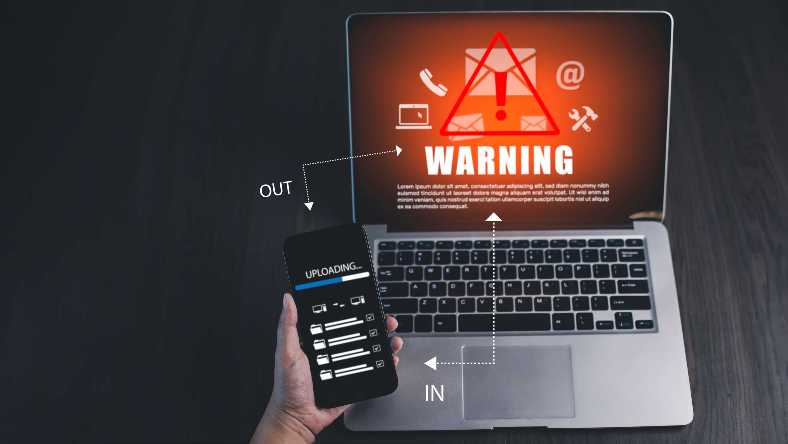 Cloud ransomware vulnerability assessment. A hand holds a smartphone above an open laptop. On the laptop screen is a vivid red warning symbol with an exclamation mark inside a triangle, along with icons for email and settings. White arrows labeled "IN" and "OUT" point between the phone and laptop, suggesting a file upload or data transfer in progress. The scene conveys a cyberattack, security breach, or phishing warning.