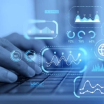 Compliance-Driven Patch Updates. Close-up of a person's hands typing on a laptop keyboard. Bright blue futuristic graphics — including graphs, pie charts, world maps, and percentages — float around the hands, suggesting data analytics, financial modeling, or business intelligence work happening in real time.