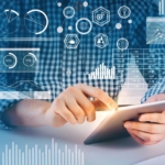 Email Phishing Defense Solutions. A person in a blue and white checkered shirt is seated at a desk, using a tablet with both hands. Floating over the image are bright digital graphics: pie charts, bar graphs, hexagonal icons, and financial data overlays. It gives the impression of someone analyzing data or using business intelligence tools in a futuristic digital workspace.