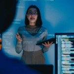 Proactive Infrastructure Monitoring. A woman stands in a dark room, illuminated by a blue glow from projected code or data on the wall behind her. She holds a tablet and appears to be giving a technical presentation. Part of a computer screen showing lines of code is visible in the foreground, slightly blurred.