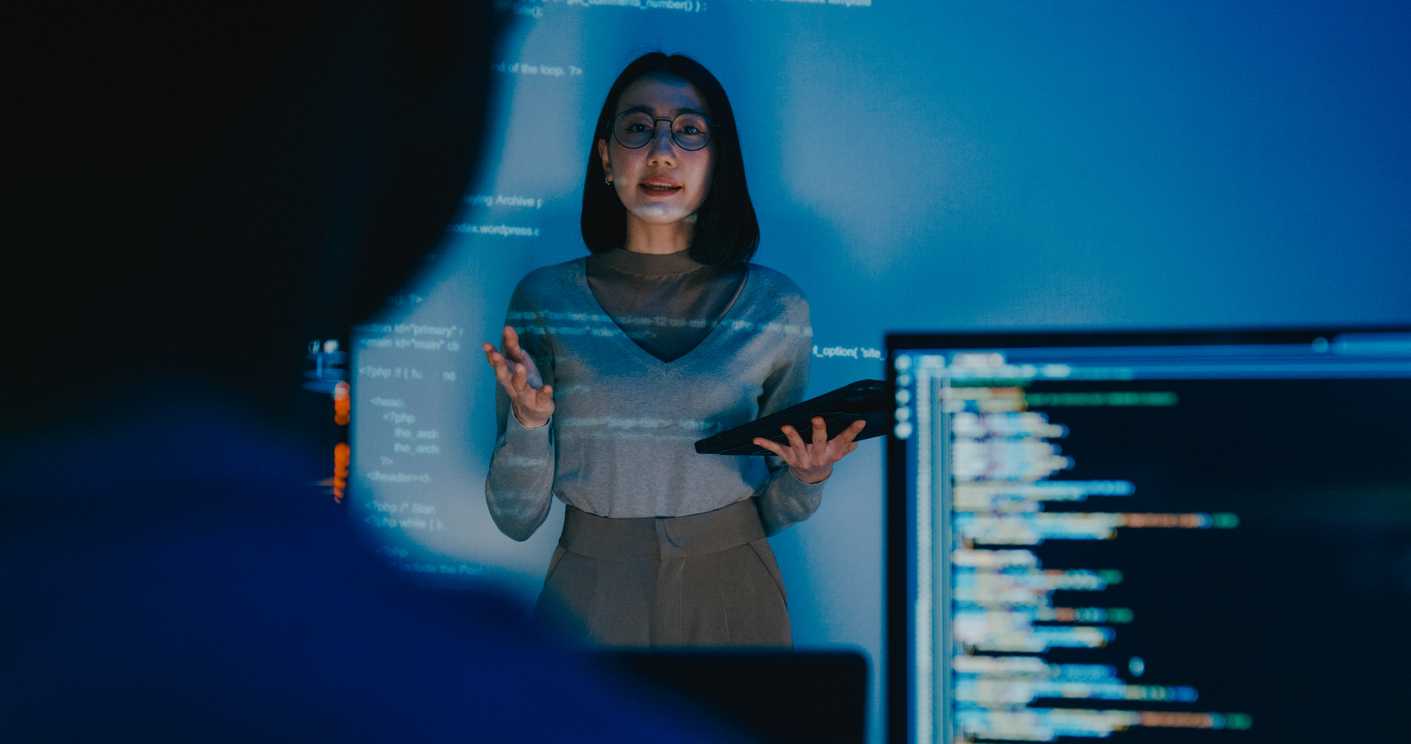 Proactive Infrastructure Monitoring. A woman stands in a dark room, illuminated by a blue glow from projected code or data on the wall behind her. She holds a tablet and appears to be giving a technical presentation. Part of a computer screen showing lines of code is visible in the foreground, slightly blurred.