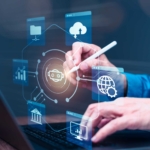 Ransomware risk management framework. A professional wearing a blue shirt is interacting with a virtual, transparent interface emerging from their laptop. They use a stylus pen, suggesting precise control. Floating around their hands are various icons symbolizing technology, including cloud services, data analytics, global connectivity, finance or banking, and artificial intelligence. The image conveys the themes of digital transformation, data analysis, and secure management through technology.