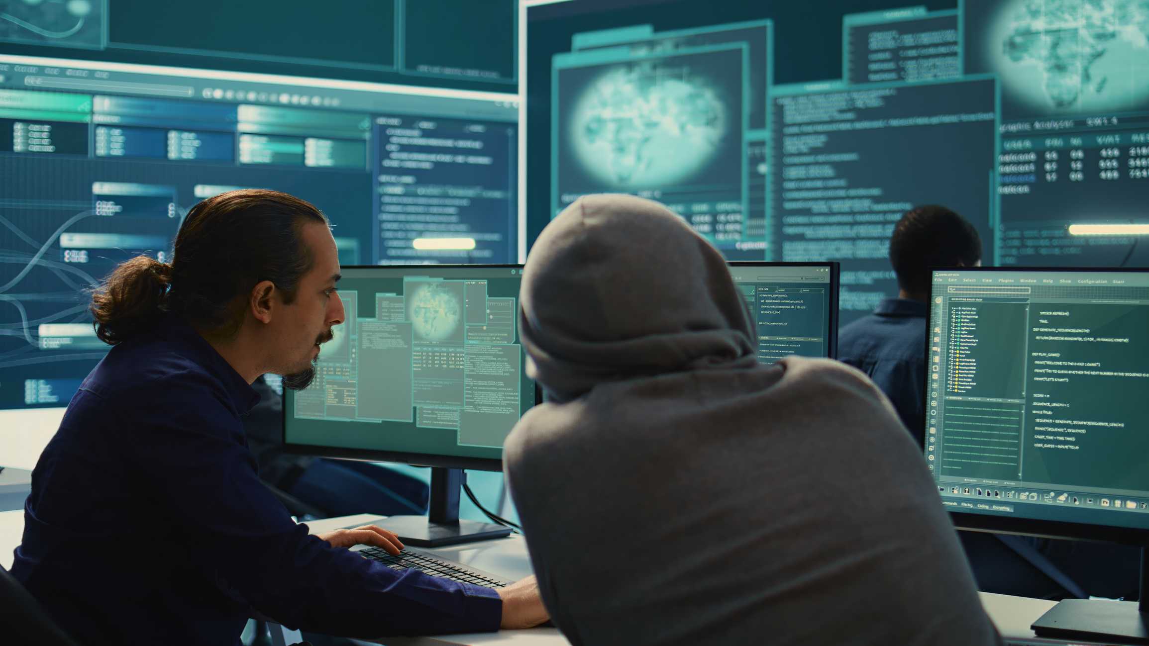 SIEM tools for ransomware detection. Two people sit side-by-side at computer monitors in a dark, high-tech room. One person, wearing a navy shirt and a ponytail, is actively typing and talking. The second person, in a gray hoodie, leans forward toward the screens. Behind them, large digital displays cover the wall, showing glowing blue-green world maps, codes, and network diagrams — suggesting they are working on cybersecurity, hacking, or network operations.