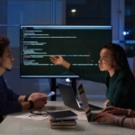 Secure Data Backup Solutions. Three colleagues sit around a table in a dimly lit office with large windows behind them showing nighttime city lights. They are engaged in a discussion in front of a large computer monitor displaying code. One woman is pointing at the screen while speaking, while another woman and a man follow along attentively with laptops and smartphones in hand. There are notebooks and glasses on the table.