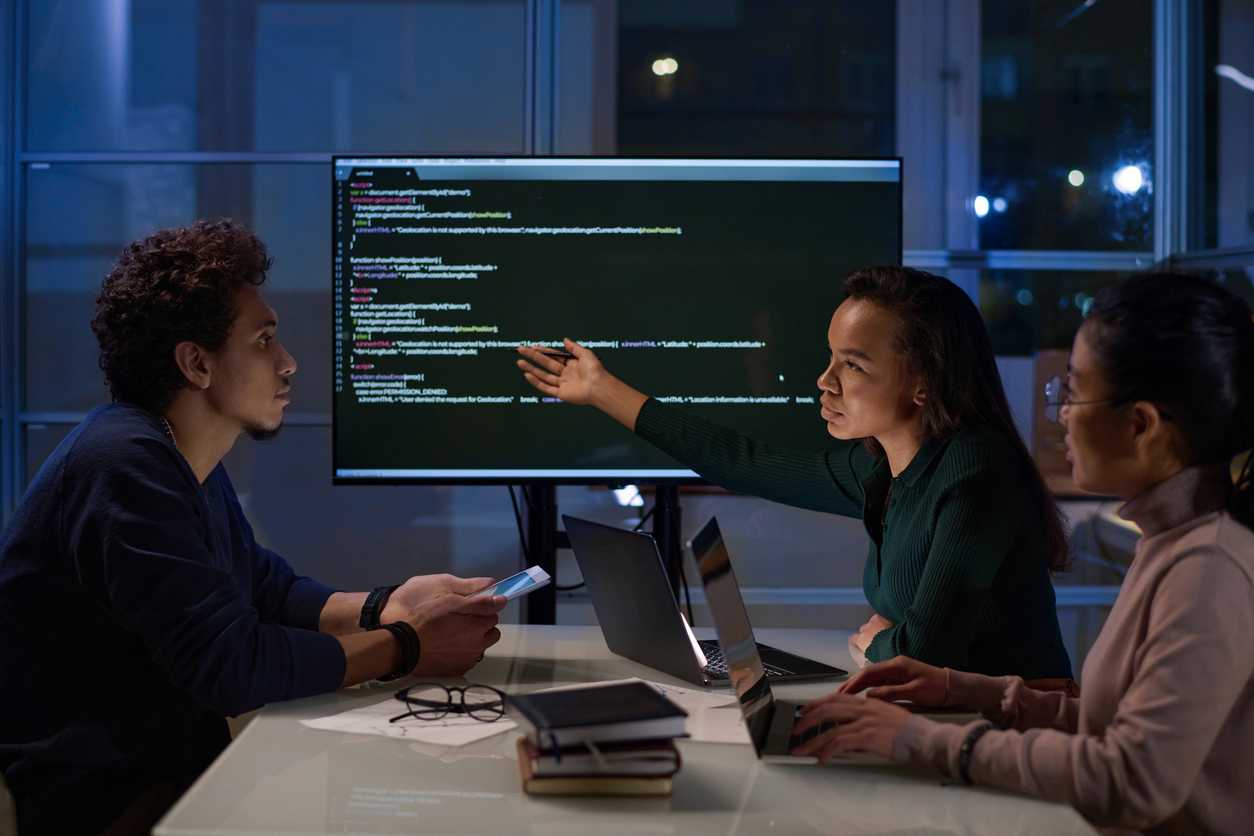 Secure Data Backup Solutions. Three colleagues sit around a table in a dimly lit office with large windows behind them showing nighttime city lights. They are engaged in a discussion in front of a large computer monitor displaying code. One woman is pointing at the screen while speaking, while another woman and a man follow along attentively with laptops and smartphones in hand. There are notebooks and glasses on the table.