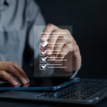 Secure Email Filtering for Businesses. A person in a gray button-down shirt uses a touchscreen device. Floating above their hand is a transparent checklist graphic with several items marked. The image suggests digital task management, compliance checking, or project organization.