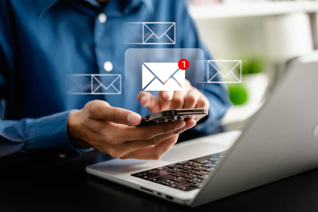 VPN ransomware protection strategies. A close-up image shows a person in a blue button-down shirt using a smartphone while sitting at a desk with a laptop open. Floating above the phone are transparent email icons, and one email has a red notification badge, symbolizing a new incoming message. The setting appears modern and professional.