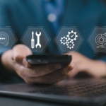 Preventative Software Patching Services. A close-up of a person using a smartphone in front of a laptop. Overlaid on the image are transparent icons representing tech support tools: a headset, screwdriver and wrench, gears with check marks, and a globe with a gear—symbolizing digital infrastructure or IT service support.