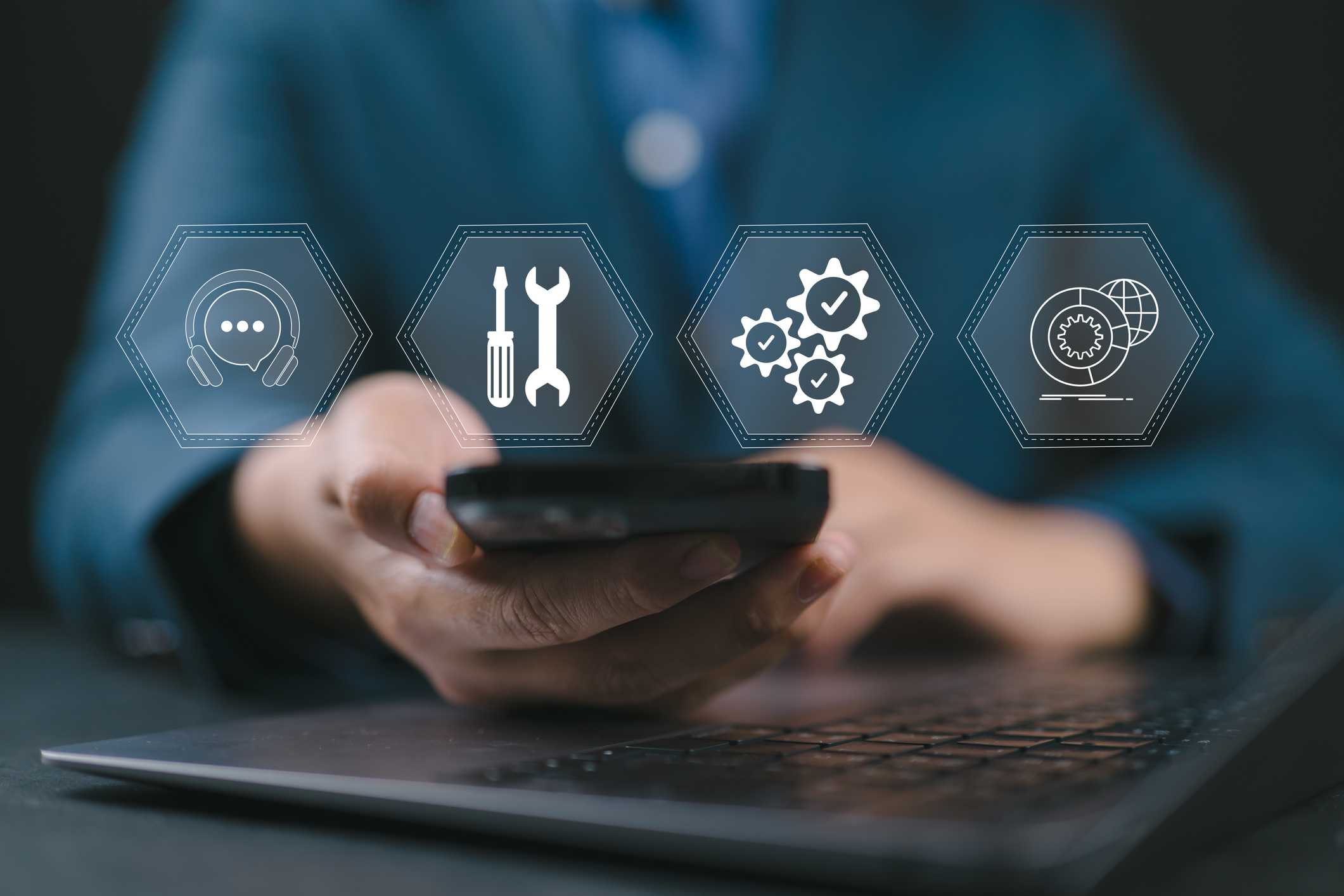 Preventative Software Patching Services. A close-up of a person using a smartphone in front of a laptop. Overlaid on the image are transparent icons representing tech support tools: a headset, screwdriver and wrench, gears with check marks, and a globe with a gear—symbolizing digital infrastructure or IT service support.
