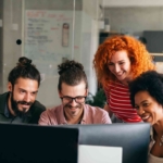 Email Threat Detection Solutions. Four colleagues huddle around a computer, laughing and smiling together, capturing a moment of friendly teamwork in an open office.