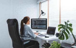 Network Traffic Analysis Services. A woman is working at a white desk in a bright home office, using a desktop and laptop with code on the screens, surrounded by green houseplants and blinds filtering in natural light.