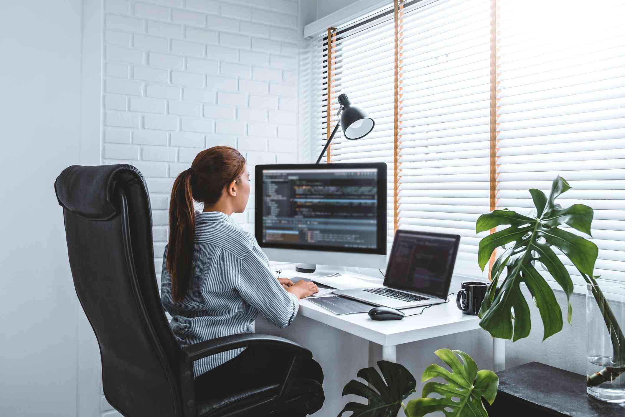 Network Traffic Analysis Services. A woman is working at a white desk in a bright home office, using a desktop and laptop with code on the screens, surrounded by green houseplants and blinds filtering in natural light.