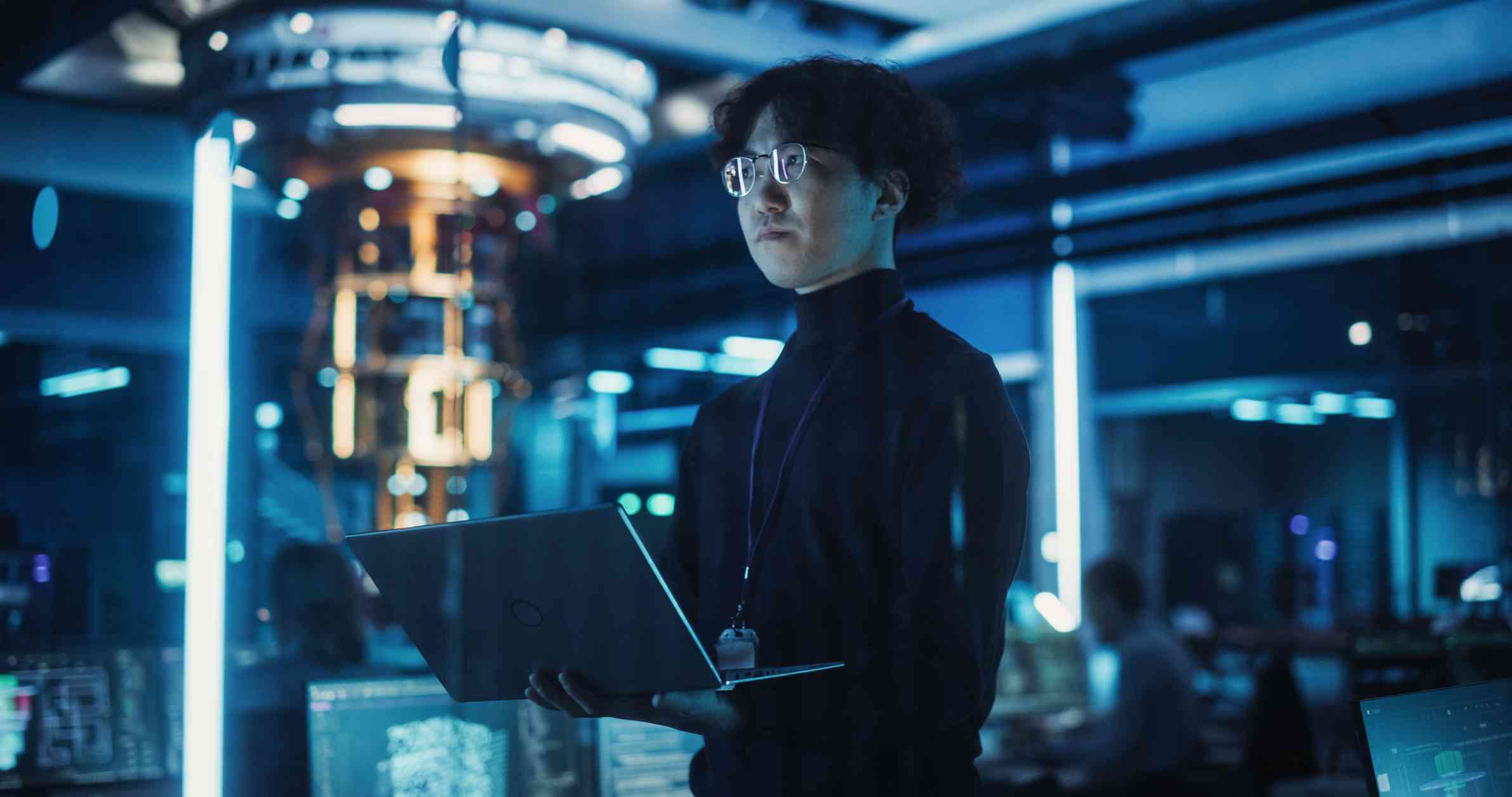 Rapid Disaster Recovery Solutions. A young person with glasses stands confidently in a high-tech data center, holding a laptop. Bright blue and white LED lights illuminate the room, casting a futuristic glow. It’s a scene of high-level tech work in a secure environment.
