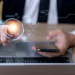 BYOD Endpoint Protection Strategies. A businessperson pointing at a glowing orange fingerprint scanner hologram while holding a smartphone, with faint futuristic interface graphics around the image.