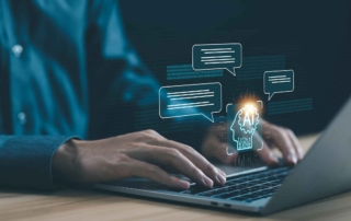 Blockchain-Based Cybersecurity Solutions. A person is typing on a laptop at a desk. The background is dark, and digital chat bubbles are hovering above the keyboard, symbolizing artificial intelligence and communication. A glowing icon of a human head with circuitry, labeled "AI," is centered above the person's hands, suggesting the use of AI tools or machine learning.
