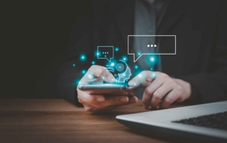 DNS Threat Filtering Services. A person in dark clothing is sitting at a wooden table, holding a smartphone in both hands. The background is softly blurred, with a laptop visible off to the side. Floating above the phone are glowing digital icons in blue, including a gear symbol with “AI” in the center, chat bubbles, and ellipsis (…). These represent artificial intelligence, chatbots, and digital messaging. The scene conveys modern communication, smart technology, and mobile-driven workflows in a business or tech setting.