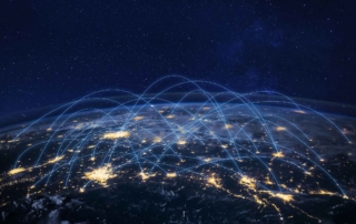 IAM Security Solutions. A satellite view of Earth at night shows city lights across the globe. Overlaid across the image are glowing blue arcs connecting various points, representing a global digital network, data exchange, or internet connectivity. The arcs give a sense of motion and interconnection between regions.