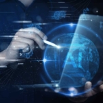 MFA Deployment Strategies. A person is using a stylus to interact with a glowing 3D digital globe. The background is a futuristic, dark interface filled with motion graphics and holographic projections, including tiny vehicles and industrial equipment. The visual suggests global digital infrastructure, smart technology, or connected industries.