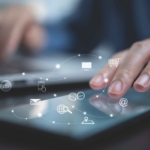 Next-Gen Antivirus Tools. A person is interacting with a touchscreen tablet, gently tapping it with their index finger. The scene is close-up and softly lit, with a blurred laptop in the background. Overlaid on the image are glowing white icons representing various digital tools and services: an envelope (email), a shopping cart, a globe, a location pin, a folder, and a mobile device. These icons are connected by dotted lines forming a network or cloud, symbolizing cloud computing, digital workflows, or interconnected systems.