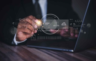 Ransomware Risk Assessments. A person in a dark suit uses a laptop on a wooden desk. Overlaid graphics show floating icons and text such as "Creative Process," cloud upload/download symbols, graphs, and chat bubbles. The person’s hand gestures as if interacting with the digital elements, conveying themes of innovation or tech development.