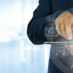 Zero Trust Network Implementation. A business professional in a suit uses a tablet device. Overlay icons are arranged in a circular interface around the device, featuring symbols for email, shopping cart, user profiles, globe, and documents. The person is tapping the screen, emphasizing business operations, mobile productivity, or cloud computing.
