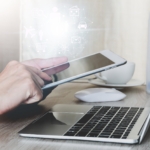 Cyber Gap Analysis Techniques. A person holding a tablet and interacting with it while sitting in front of a laptop. The devices are on a wooden desk, and a white coffee cup is nearby. The overall tone is warm and soft, suggesting a cozy remote work or hybrid work setting.