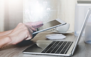 Cyber Gap Analysis Techniques. A person holding a tablet and interacting with it while sitting in front of a laptop. The devices are on a wooden desk, and a white coffee cup is nearby. The overall tone is warm and soft, suggesting a cozy remote work or hybrid work setting.