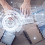 Ransomware recovery communication protocol. A top-down shot of someone typing on a white keyboard surrounded by notebooks, papers, a coffee cup, and eyeglasses. A translucent circular HUD (heads-up display) graphic overlays the scene, symbolizing data entry or secure documentation.